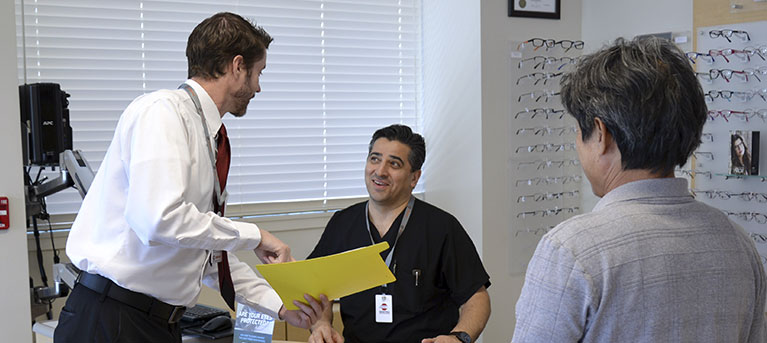 An image of an eye doctor in a white dress shirt and tie going over a medical chart with a lab technician while in blue scrubs with a patient onlooking in the foreground. All are in an examination room.